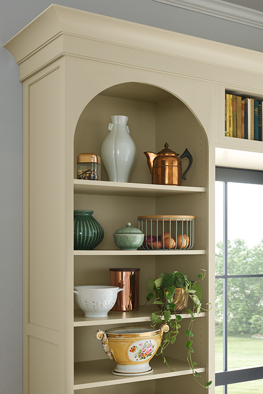 This is a built-in kitchen shelf, styled as a bookshelf, with items such as vases, collinders, plants, and produce on display. 