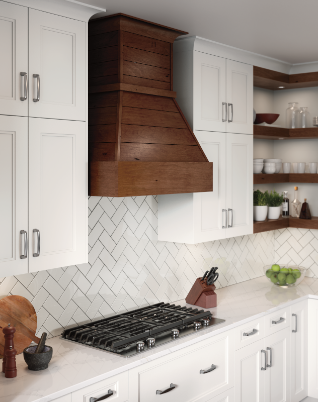 This kitchen close-up includes white cabinetry, a natural wood-toned range hood, matching wood-toned shelves, and a minimalist white tile backsplash that creates visual intrigue. 