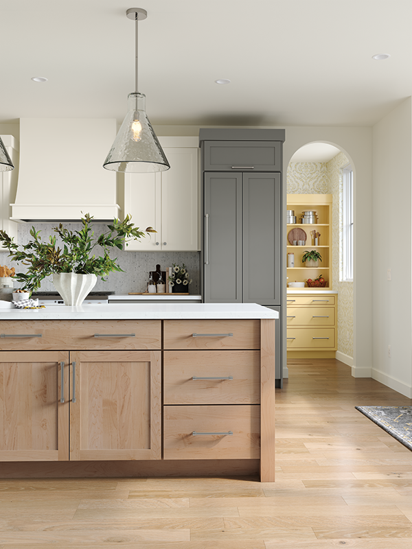 In this photo is a kitchen island in the foreground, comprised of natural wood-toned cabinets and marble countertop. In the background is a paneled refrigerator that complements the design of the kitchen, concealed in grey-colored paneling to appear as cabinetry. 