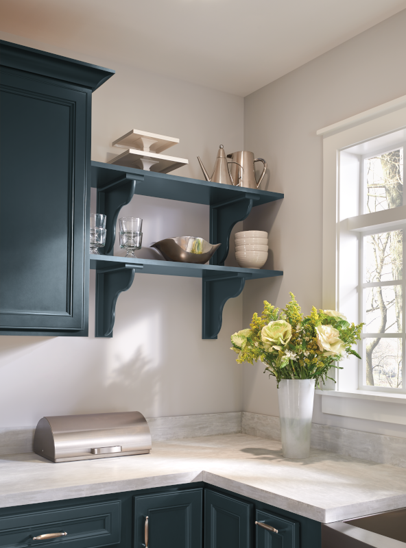 The focus of this image is a pair of kitchen shelves, with corbel-style detailing that curves from the wall up to the shelf to add aesthetic detail as well as shelf support. The shelves and corbels are both painted a deep chambray blue. Kitchen items are on display, with cups and bowls on the shelves, and a lovely floral arrangement of greenery on the kitchen counter.