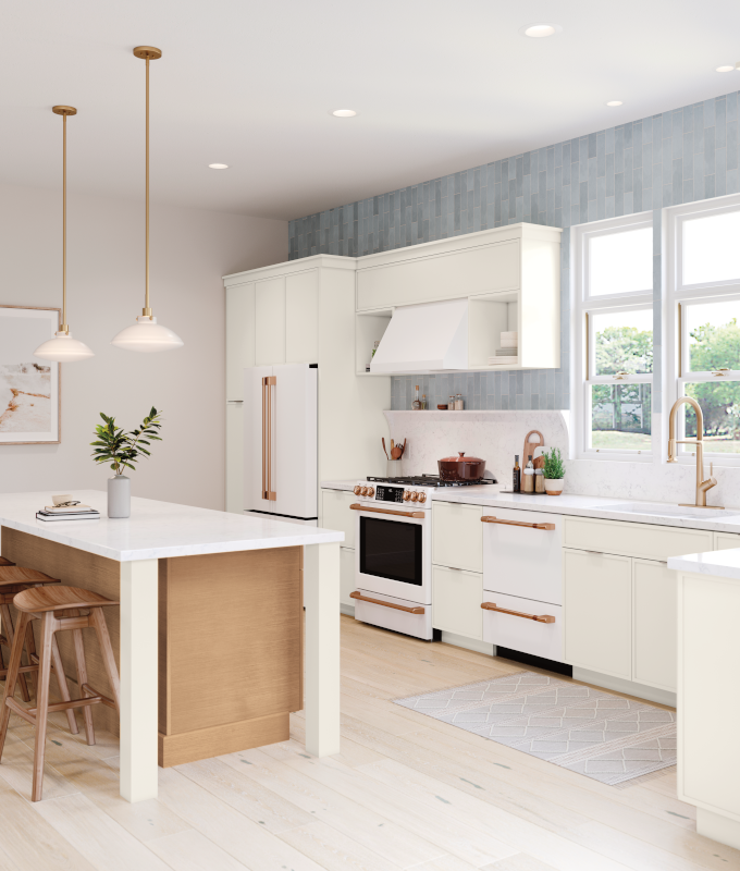 A modern kitchen design boasting off-white lower and upper cabinetry, white fridge with gold hardware, matching white appliances, white range hood, and rose gold-colored faucet. 