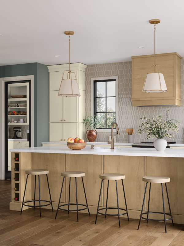 A natural-toned kitchen embraces light wooden details on the sides of the kitchen island and the oven range hood, an off-white cabinet installation to the left, and marble countertops. The counter is set with a bowl of fruit, an arrangement of greenery, and a few kitchen essentials for a put-together but lived-in feel. A textured backsplash of white and taupe ties the entire space together. 