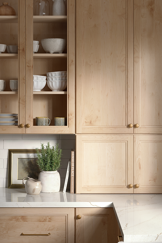 The image is a close-up of an ivory-colored marble countertop set with two books, a plant, and a framed picture, with cabinets above and below. These light-colored natural wood cabinets show a section to the left with glass-front doors - a detail that displays cups and bowls inside. 