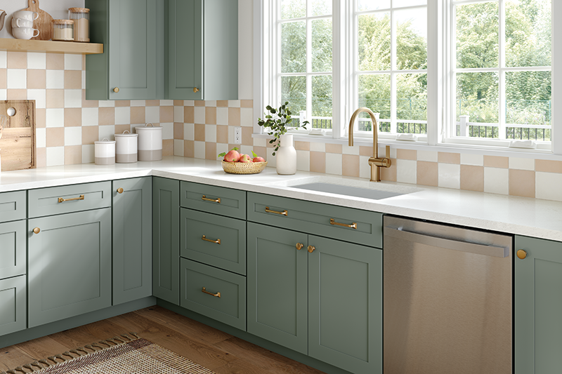 This kitchen corner is uniquely detailed in taupe and ivory checkered tile backsplash. The cabinets are a seafoam green color, and the overall feel is modern, with brass accents and neutral-colored pots and bowls. At the top right are three white-paned windows behind the sink with a view of the trees in the yard. 