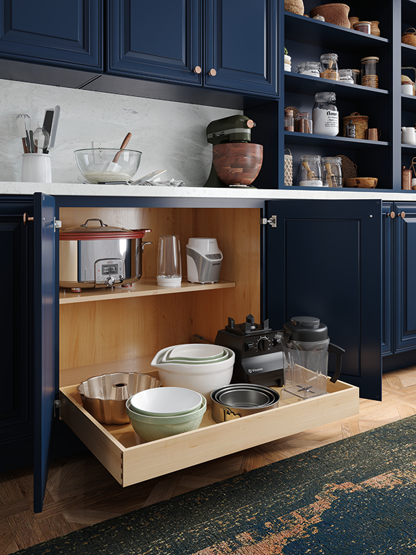 A dark blue lower kitchen cabinet, opened, with drawer storage and shelves storing bowls and appliances. 
