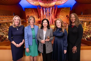 Five supervisors stand side by side in a brightly lit concert hall with colorful patterned seating and a large abstract wooden structure rising behind them. From left to right, Supervisor Kathryn Barger, Supervisor Janice Hahn, Supervisor Hilda Solis (Chair), Supervisor Lindsey Horvath, Supervisor Holly Mitchell. They wear formal attire including dresses, a blazer over a green dress, a checked blazer with a gavel in hand, a navy satin dress, and a black dress with a red necklace that reads 'love'; warm lighting highlights the wooden architectural details and curved ceiling panels throughout the hall.