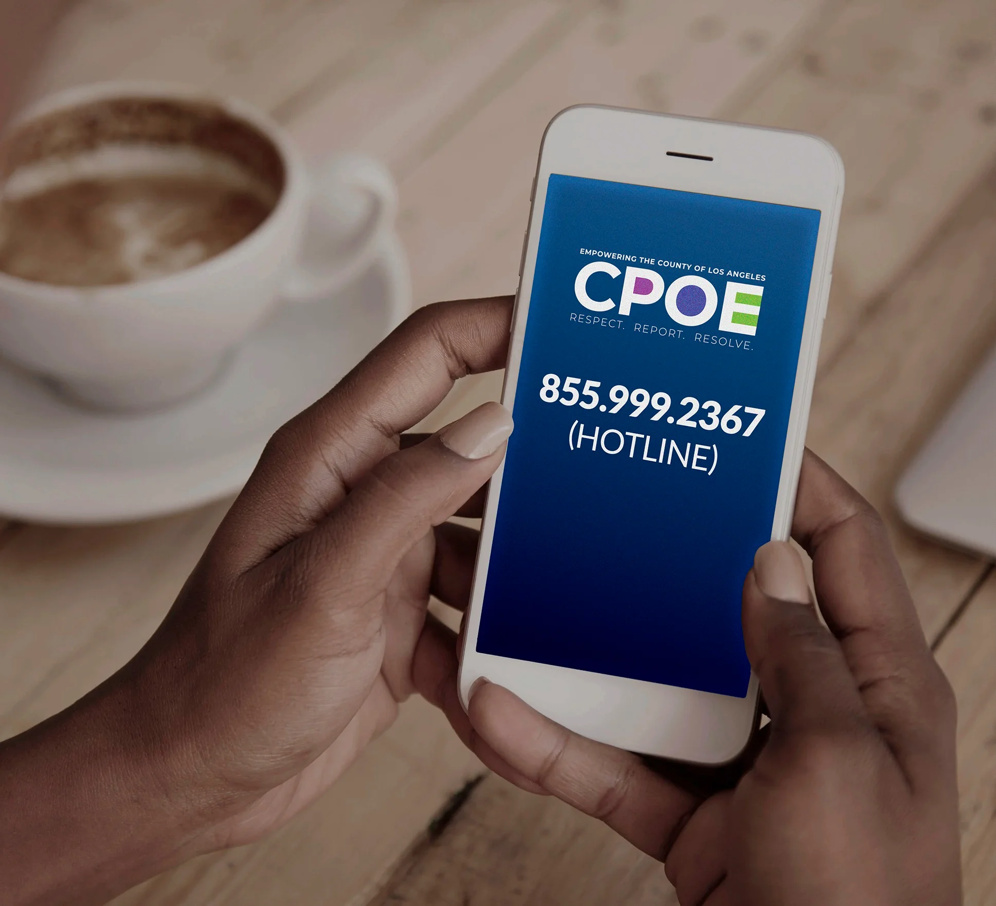 A person holds a smartphone displaying a blue screen with the text 'CPOE' and '855.999.2367 (Hotline)' in white lettering; the phone is held above a wooden table with a white cup of coffee and saucer in the background.