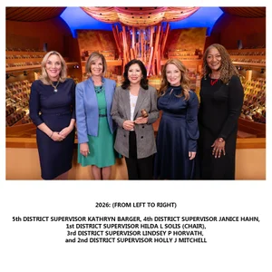 Five supervisors stand together in a brightly lit concert hall with colorful patterned seating and a large abstract wooden structure rising behind them; they wear formal attire including a navy dress, a green dress with a blue blazer, a checked blazer while holding a gavel, a navy satin dress, and a black dress with a red necklace; below the photo is centered black text reading '2026: (From Left to Right) 5th District Supervisor Kathryn Barger, 4th District Supervisor Janice Hahn, 1st District Supervisor Hilda L Solis (Chair), 3rd District Supervisor Lindsey P Horvath, and 2nd District Supervisor Holly J Mitchell'.