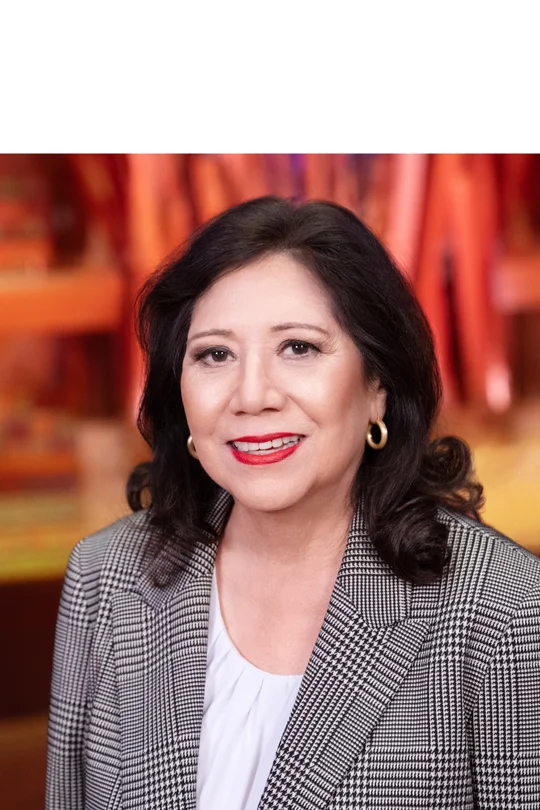 Supervisor Solis with long dark hair and gold hoop earrings smiles while wearing a black-and-white houndstooth blazer over a white blouse; the background is softly blurred with warm orange and red tones and abstract vertical shapes.