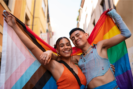 Two people stand close together smiling and posing in a narrow outdoor street lined with buildings, each holding a pride flag behind their shoulders; the person on the left holds a Progress Pride flag with a chevron of black, brown, light blue, pink, and white stripes layered over a rainbow background, while the person on the right holds a rainbow pride flag with horizontal red, orange, yellow, green, blue, and purple stripes; both wear sleeveless tops and jewelry, with arms raised and bodies angled toward each other, conveying a celebratory and joyful moment.