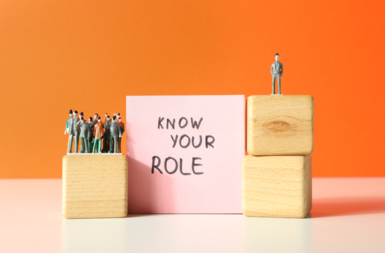 Small figurines of people stand on stacked wooden blocks on both sides of a pink card with text that reads KNOW YOUR ROLE. A group of figurines stands together on a single block on the left, while a single figurine stands on two stacked blocks on the right. The background is a solid orange color with a light surface beneath the objects.
