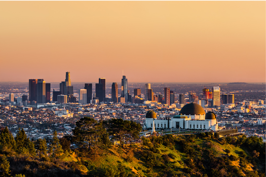 A wide panoramic view shows the Los Angeles skyline at golden hour with downtown skyscrapers rising across the horizon; in the foreground, the white domed Griffith Observatory sits atop a green hillside with trees and walking paths; the city below spreads out with dense neighborhoods and streets, and the sky above is a warm orange gradient fading into light blue near the horizon.