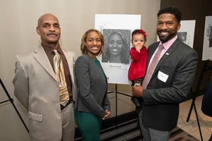 4 people posing for a photo in front of another woman's photo.
