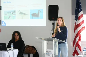 A group of people sitting at tables at an event while a woman speaks to them.