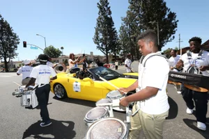 Supervisor Mitchell holding up drumsticks while in a car in the middle of a circle of drummers