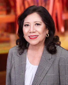Supervisor Solis with long dark hair and gold hoop earrings smiles while wearing a black-and-white houndstooth blazer over a white blouse; the background is softly blurred with warm orange and red tones and abstract vertical shapes.