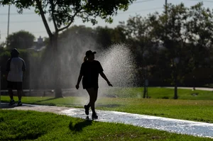 a woman walking down the sidewalk with grass and sprinklers around her.
