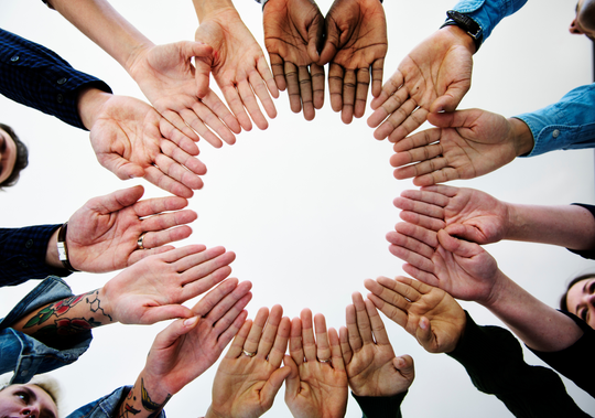 A circle of multiple people’s open hands reaching inward toward the center, palms facing up, forming a ring. The hands vary in skin tone and appear from different directions around the frame against a light background. Some wrists show clothing sleeves, watches, or tattoos.