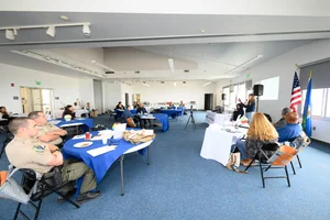 A group of people sitting at tables at an event while a woman speaks to them.