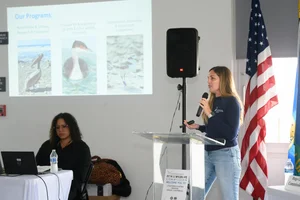 A group of people sitting at tables at an event while a woman speaks to them.