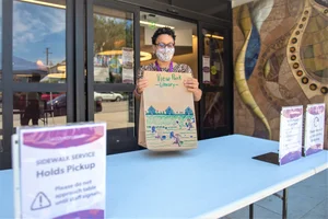 Woman in a mask at a table holding up a "View Park Library" sign.