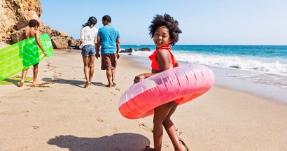 Family of four enjoying a day at the beach, walking with inflatable toys. Find term life insurance.