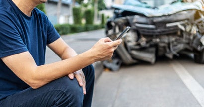 Man using phone to call his insurance agent after traffic accident with a background of the crashed car. How to file Car Insurance Claims.