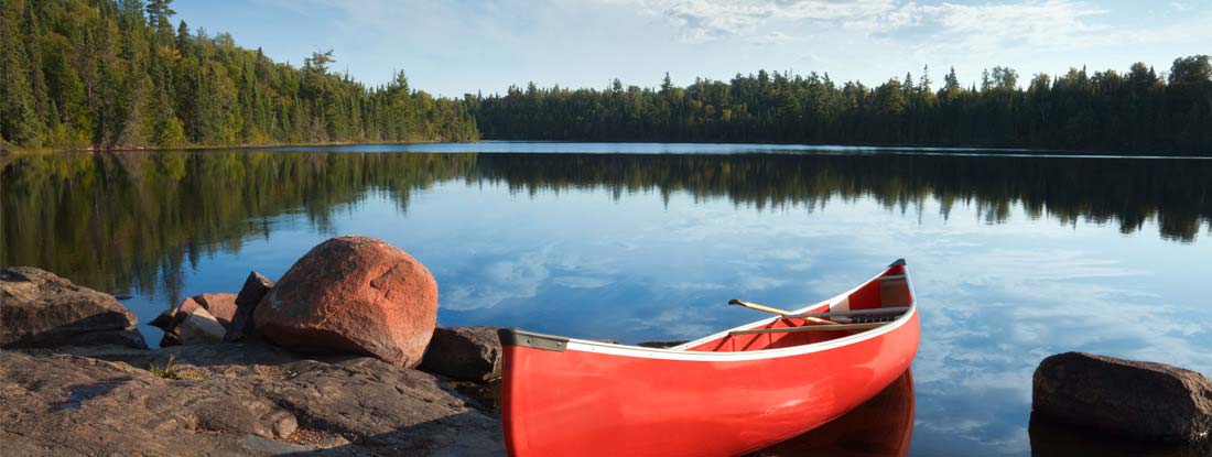 Red canoe on rocky shore of calm northern lake. Find Canoe Insurance.