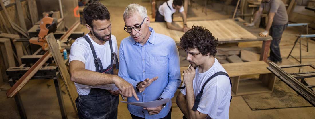 Manager and manual workers reading design plans in a workshop. What Is a Minimum Premium Workers' Compensation Policy?