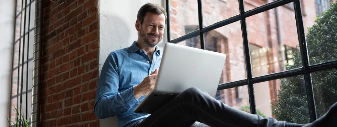 Man sitting on window sill, using laptop. Cheapest Renters Insurance Companies of 2026.