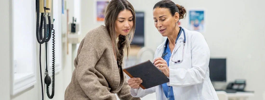 Young female sits up on an exam table during a routine medical appointment with her doctor. Find Health Insurance.