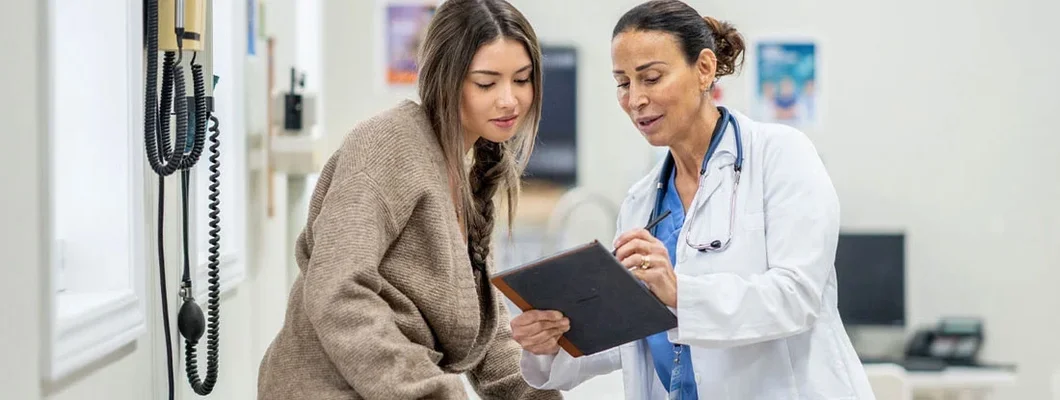 Young female sits up on an exam table during a routine medical appointment with her doctor. Find Health Insurance.