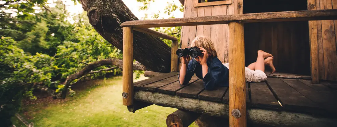 Boy in a treehouse looking in the distance with binoculars. Why Backyard Fun Isn’t All Fun and Games.