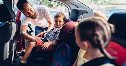 Father putting his kids in carseats to be safe during road trip. The 10 Safest Cars.