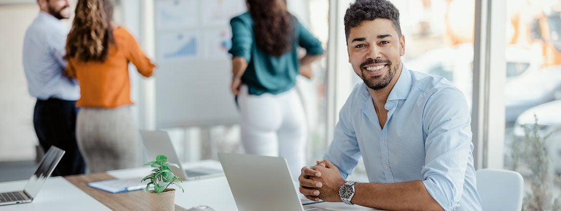 Smiling working man looking at camera with colleagues in the background. Find Business Umbrella Insurance. 