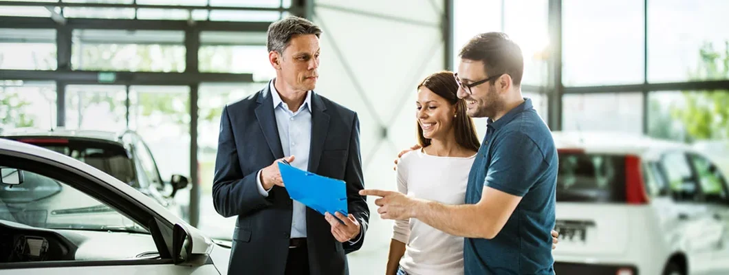 Couple having a conversation with a salesperson at a car dealership, all of them standing next to new cars. Find Car Dealer Insurance.