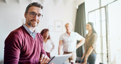 Businessman looking at the camera while working on his tablet and sitting in the office with employees in background.Find group disability insurance.