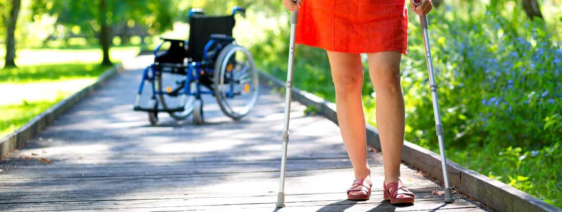 Woman practicing walking on crutches in park and wheelchair in the background. How Long Does Group Long Term Disability Last?