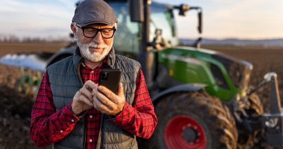 Senior farmer with mobile phone in field with tractor in background. Helpful Tips for Buying New or Used Farm Equipment. 