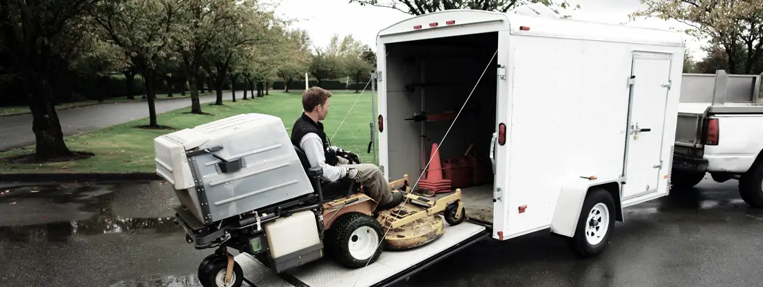 A man is loading a zero-turn lawn mower into a white enclosed cargo trailer.  Find Cargo Trailer Insurance.