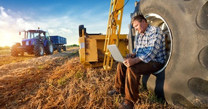 Farmer using a laptop in a field next to a tractor and other farm equipment. How much does farm insurance cost.