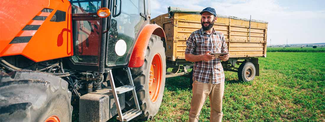 Farmer with a digital tablet standing next to a tractor and a trailer in a field. What every farmer needs to know about insurance.