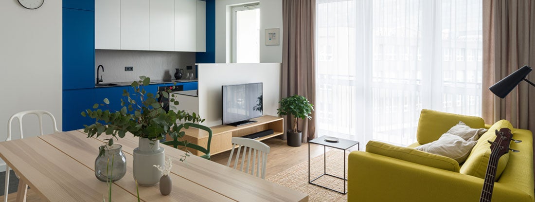 Open modern apartment with wooden dining table, different chairs with small, blue kitchen open to living room with yellow couch and tv. Find Montana renters insurance.
