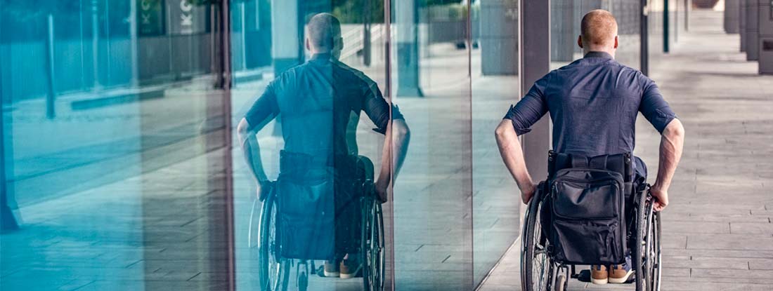 Man in a manual wheelchair outdoors on a paved area next to a modern building with large glass windows. Find Long Term Voluntary Cost Insurance.