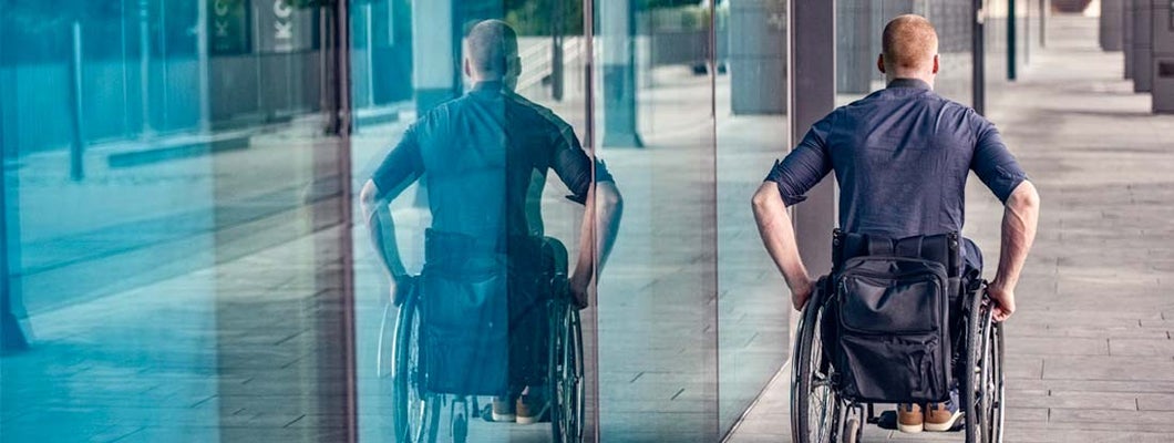 Man in a manual wheelchair outdoors on a paved area next to a modern building with large glass windows. Find Long Term Voluntary Cost Insurance.