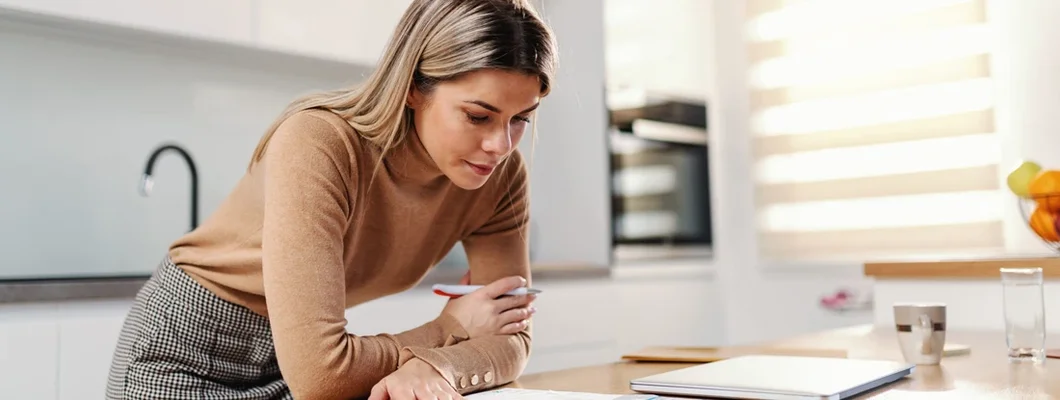 Businesswoman leaning on kitchen table and filling up bills while standing in kitchen at home. Find Business Insurance in Chevy Chase, Maryland. 