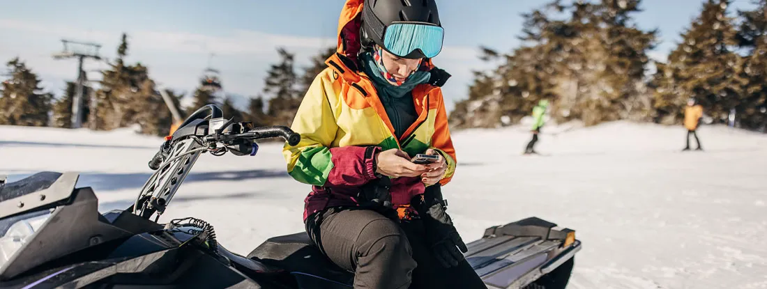 Woman sitting on snowmobile on hill of a snowcapped mountain using smart phone. Should I Get a GPS for My Snowmobile?