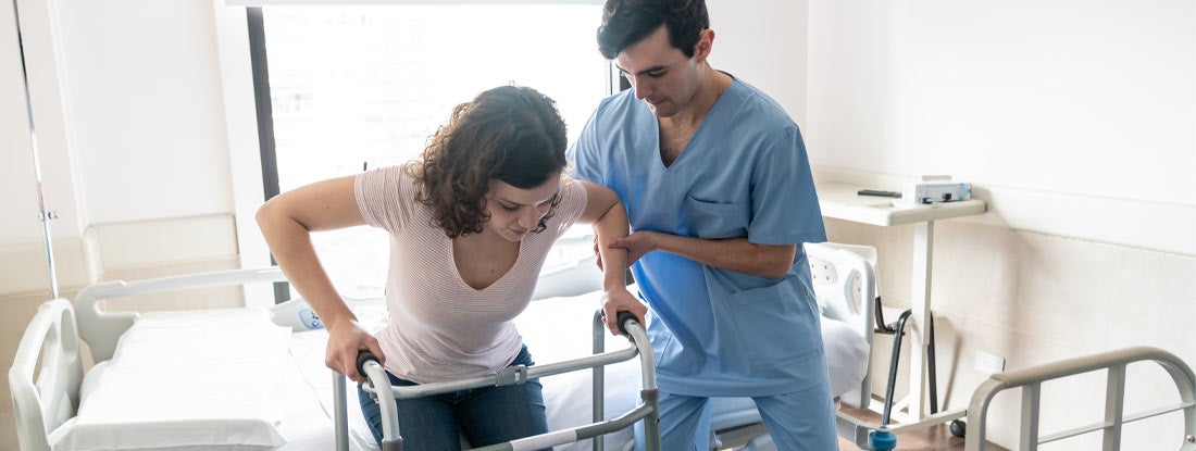 Nurse helping a patient stand up from hospital bed while she leans on walker. Workers' Compensation Medical Coverage.