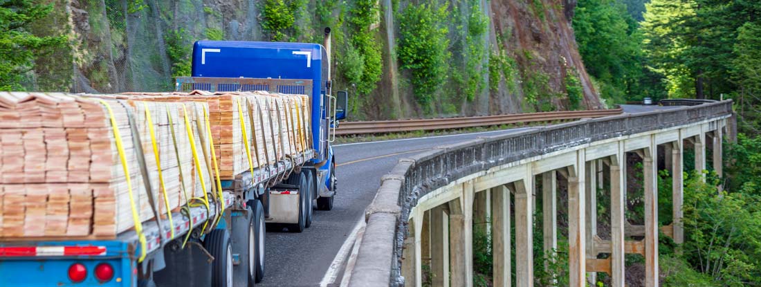 Semi truck transporting lumber wood on commercial flatbed semi trailer driving on the mountain bridge in Columbia Gorge. Find commercial trailer insurance.