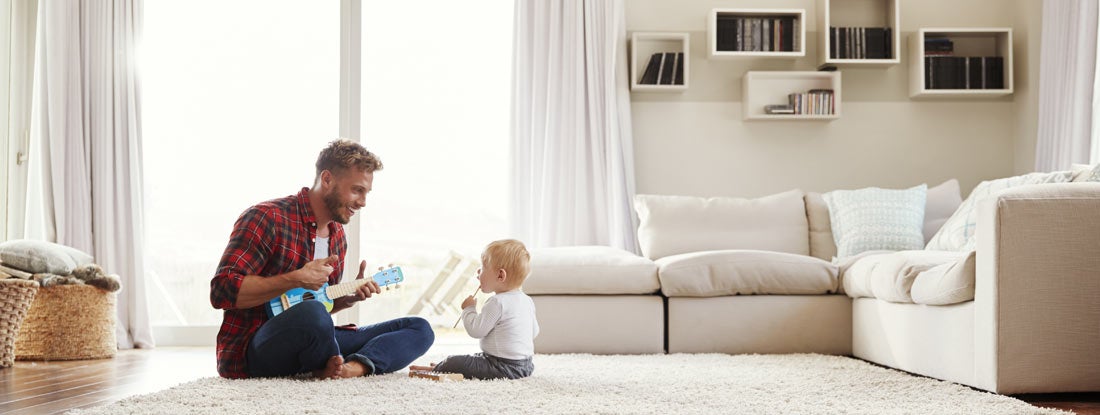 Father playing ukulele with young son in their sitting room. Find Universal Life Insurance. 